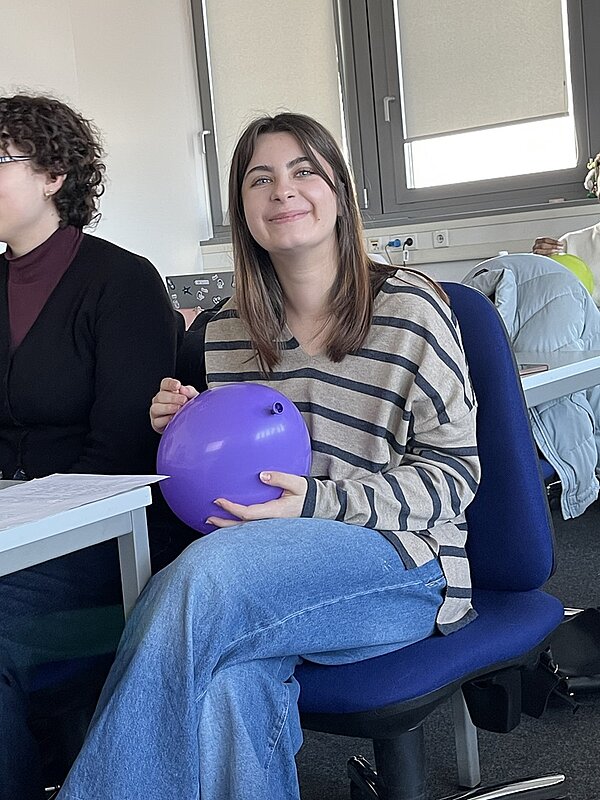 Eine Auszubildende mit schulterlangem braunem Haar sitzt auf einem blauen Stuhl und hält einen lilafarbenen Luftballon in den Händen. Sie trägt ein gestreiftes, langärmliges Oberteil und eine hellblaue Jeans. Im Hintergrund sind ein Fenster, ein weißer Heizkörper und eine hellblaue Jacke auf einem weiteren Stuhl zu sehen. Neben der Person sitzt eine weitere Person mit lockigem, dunklem Haar und Brille, die ein dunkles Oberteil und einen schwarzen Blazer trägt. Auf dem Tisch liegen Papiere.