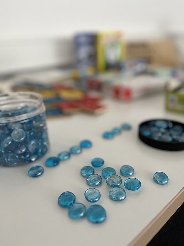 Glas mit blauen Glassteinen, die locker über einen Tisch verteilt sind. Im Hintergrund unscharf weitere Lernmaterialien und Spiele.