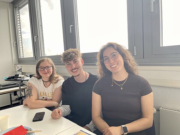 Drei Absolventinnen und Absolventen sitzen lächelnd nebeneinander an einem weißen Tisch in einem hellen Raum mit großen Fenstern. Die linke Person trägt ein weißes T-Shirt mit Aufdruck, die mittlere Person ein dunkles Oberteil, und die rechte Person ein schwarzes T-Shirt sowie eine Halskette. Auf dem Tisch liegen ein Smartphone, ein Becher und einige weitere Gegenstände. Im Hintergrund sind Tische, Stühle und eine Heizung zu sehen.