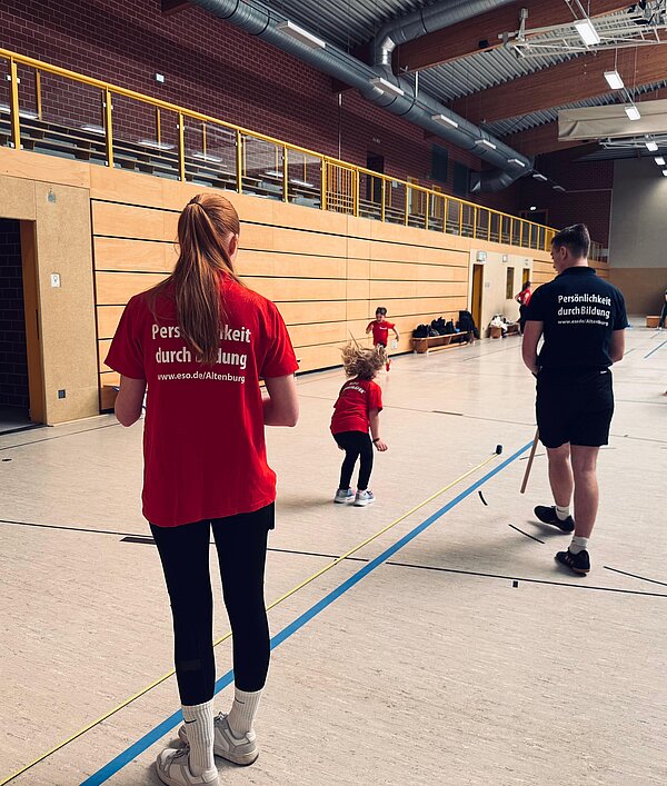 In einer Sporthalle stehen zwei betreuende Personen in roten und schwarzen T-Shirts, während ein kleines Kind in der Mitte läuft; auf den Shirts steht „Persönlichkeit durch Bildung“.