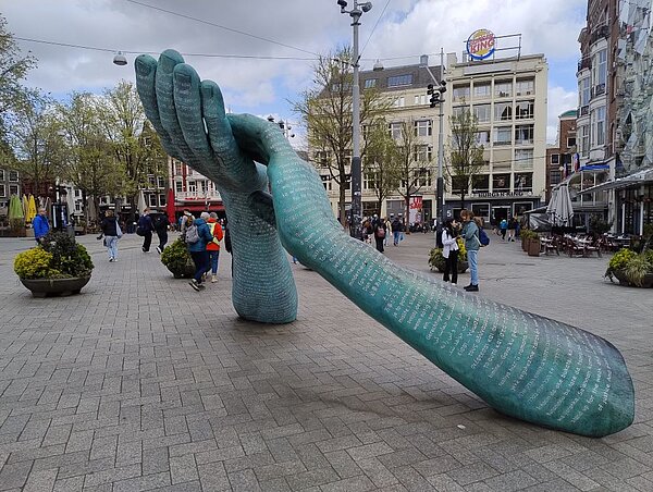 Ein ungewöhnliches Kunstwerk in einer Fußgängerzone: Eine große, stilisierte Hand, die auf dem Boden liegt. Die Skulptur ist mit Text bedeckt, und Passanten gehen vorbei.