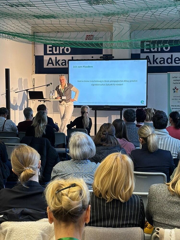 Eine Referentin steht auf einer Bühne vor einem Publikum und hält einen Vortrag zum Thema 'Zeit zum Plaudern'. Auf der Leinwand hinter ihr steht die Frage: 'Welche kleine Entscheidung in Ihrem pädagogischen Alltag gestaltet eigentlich schon Zukunft für Kinder?'. Die Veranstaltung findet im Rahmen der Euro Akademie statt, was durch die Banner im Hintergrund erkennbar ist. Das Publikum sitzt in Stuhlreihen und folgt aufmerksam dem Vortrag.
