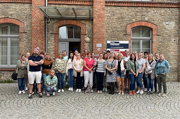 Erfolgreiche Fortbildung der Praxisanleiter*innen Eine Gruppe von Personen steht vor einem Backsteingebäude mit großen Fenstern und einem bogenförmigen Eingang. Ein Schild neben der Tür trägt die Aufschrift “Euro Akademie".”.