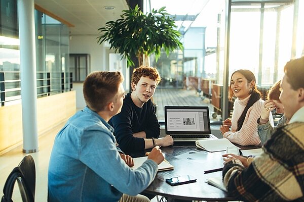 Junge Schülerinnen und Schüler, die auf dem Campus mit Büchern und Laptop um den Tisch sitzen, tauschen sich angeregt aus. 