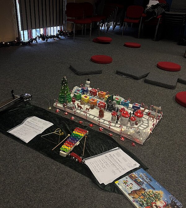 Weihnachtsleseraum mit Teppichboden: In der Mitte stehen eine leuchtende Mini-Winterlandschaft, Xylophon, Triangel und Notenblätter im Stuhlkreis.