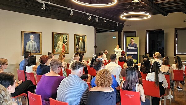 Blick in den Saal des Stadtmuseums Halle, auf das Publikum, die Absolvent*innen und auf die Bühne, auf der die Schulleiterin eine Rede hält.