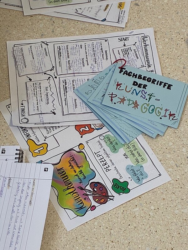 Auf einem hellen Tisch liegen mehrere Arbeitsblätter und Karteikarten, die sich mit kunstpädagogischen Fachbegriffen beschäftigen. Die Karteikarten sind hellblau und tragen handschriftlich den Titel 'Fachbegriffe der Kunstpädagogik' in bunten, teils künstlerisch gestalteten Buchstaben. Die Arbeitsblätter sind farbig gestaltet und enthalten handschriftliche Notizen, Zeichnungen und farbige Markierungen. Ein Arbeitsblatt zeigt eine bunte Farbpalette mit dem Wort 'Kunstpädagogik' in geschwungener Schrift, daneben sind weitere Begriffe und kurze Erklärungen zu sehen. Ein anderes Blatt ist als Spielfeld mit Start- und Zielpunkt gestaltet und enthält verschiedene Textfelder mit Informationen. Die Materialien wirken handgemacht und dienen offenbar der Vermittlung von Wissen im Bereich der frühkindlichen Kunstpädagogik. Die Szene entstand im Rahmen einer Exkursion ins Kunstmuseum „Kloster Unser Lieben Frauen“ in Magdeburg und zeigt die Lernunterlagen von Auszubildenden zu Erziehern.