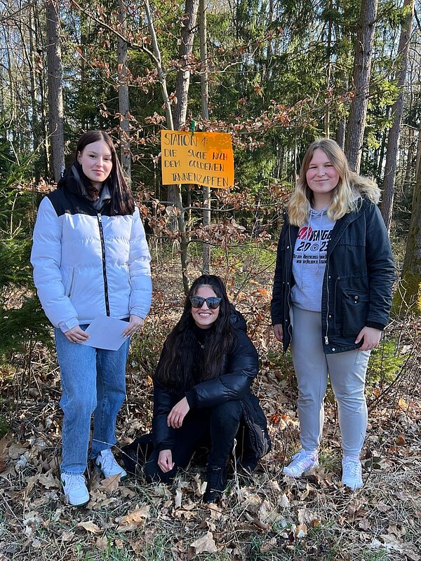 Drei junge Frauen stehen oder hocken im Wald vor einem gelben Schild mit der Aufschrift: "Station 1: Die Suche nach dem goldenen Tannenzapfen". Sie lächeln.