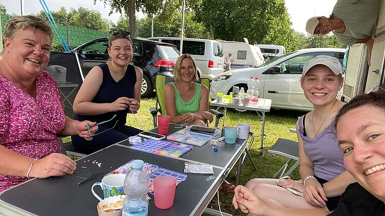 Ehemaligentreffen Fünf Personen sitzen an einem Campingtisch und lächeln. Auf dem Tisch stehen Tassen und liegen Bastelutensilien, die von den Personen gerade benutzt werden. Im Hintergrund sind Autos und Wohnwagen zu sehen.