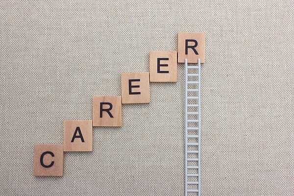 Auf einem beigen gewebten Untergrund liegen viereckige Plättchen aus Holz, angeordnet wie Treppenstufen nach oben, auf jedem der Holzplättchen stehen Buchstaben die das Wort Career ergeben, am letzten Buchstaben R, der am höchsten steht, ist eine kleine Leiter platziert, die die Karriereleiter nach oben symbolisiert.