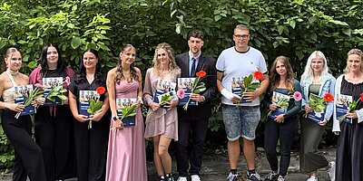 Eine Gruppe von zehn Personen steht in einer Reihe vor Büschen mit grünem Blattwerk. Sie halten jeweils eine Mappe der Euro Akademie und eine Blume in den Händen. Die Gruppe besteht aus Frauen und Männern in verschiedenen Outfits, von elegant bis lässig, und wirkt feierlich oder stolz, möglicherweise bei einer Abschlussfeier oder einem besonderen Anlass.