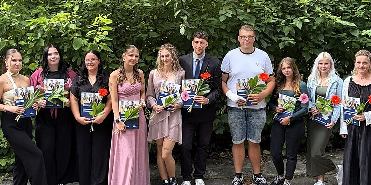 Eine Gruppe von zehn Personen steht in einer Reihe vor Büschen mit grünem Blattwerk. Sie halten jeweils eine Mappe der Euro Akademie und eine Blume in den Händen. Die Gruppe besteht aus Frauen und Männern in verschiedenen Outfits, von elegant bis lässig, und wirkt feierlich oder stolz, möglicherweise bei einer Abschlussfeier oder einem besonderen Anlass.