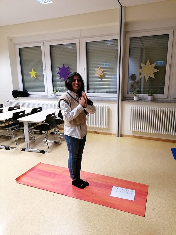 Engagiert und kreativ Eine Auszubildende steht lächelnd auf einer roten Yoga-Matte und presst die Hände zusammen.