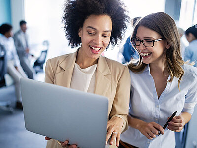 Zwei lächelnde Frauen stehen nebeneinander und schauen gemeinsam auf einen Laptop. Die Frau links hat lockige Haare und trägt ein hellbeiges Blazer-Jacket über einem weißen Oberteil. Die Frau rechts trägt eine Brille, ein hellblaues Hemd und hält einen Stift in der Hand. Beide wirken konzentriert und zufrieden. Im Hintergrund sind unscharf weitere Personen in einem Büroumfeld zu sehen. Der Raum ist hell und modern eingerichtet.