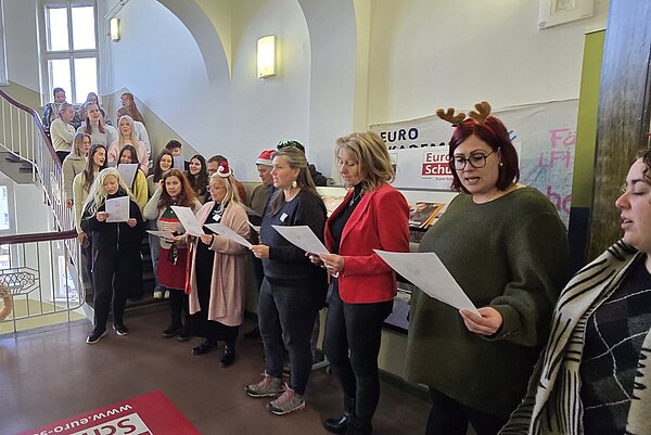 Eine große Gruppe von Lehrkräften und Studierenden steht im Treppenhaus der Euro Akademie, viele mit Weihnachtsaccessoires, und singt gemeinsam mit Notenblättern in der Hand.