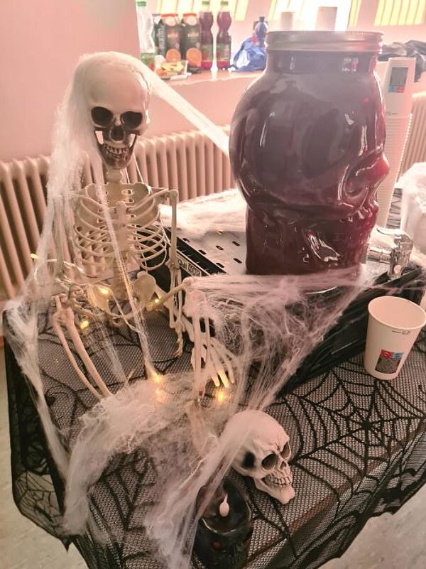 Ein dekorierter Tisch für eine Halloweenfeier mit einem Deko-Skelett, Spinnweben und einem großen mit blutrotem Saft gefüllten Glasbehälter, das wie ein Totenkopf aussieht.
