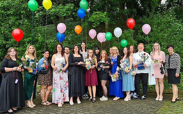 Eine Gruppe junger Frauen und zwei Männern steht festlich gekleidet vor einer grünen Wand aus Büschen und Bäumen. Bis auf die Frau rechts, die eine Lehrerin sein könnte, halten alle bunte Luftballons in der Hand, die über ihnen schweben.