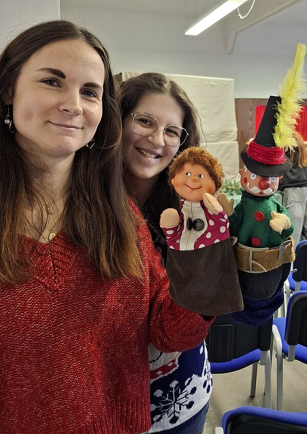 Zwei lächelnde junge Frauen halten bunte Handpuppen in einer festlich geschmückten Umgebung.