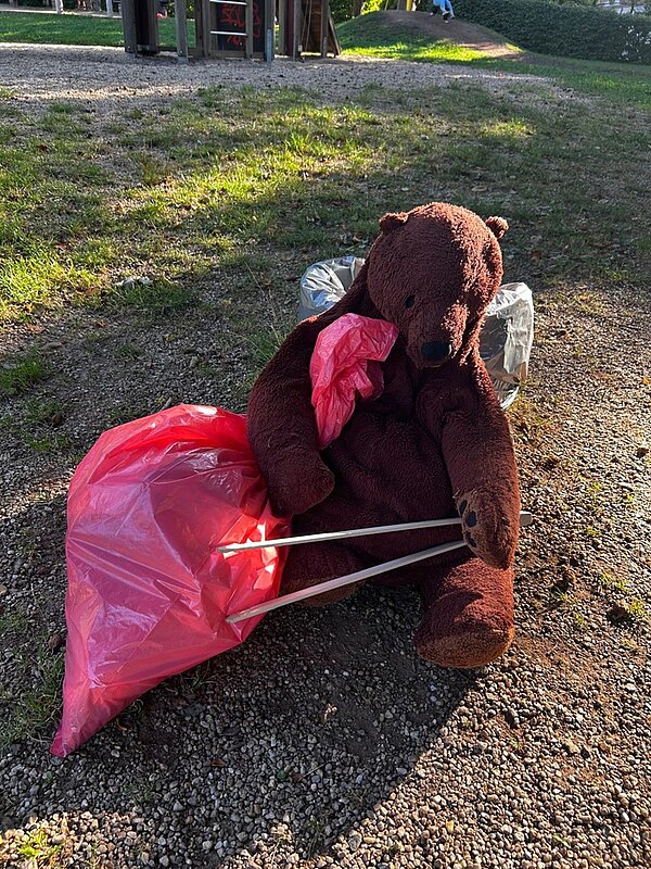 Ein großer brauner Plüschteddybär sitzt auf dem Waldboden und hat die von den Schülerinnen und Schülern gesammelten roten Mülltüten um sich herum drappiert.