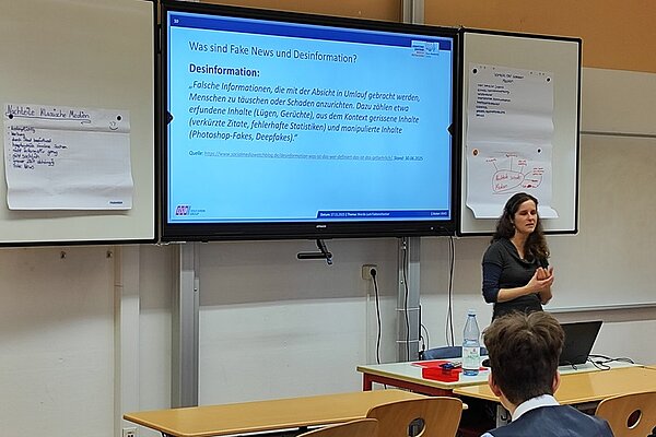 In einem modernen Seminarraum steht eine weibliche Person vor einer Präsentationsfläche und hält einen Vortrag zum Thema Desinformation im Rahmen des Projekts „Werde zum Faktenchecker“. Im Hintergrund ist ein großer Bildschirm zu sehen, auf dem eine Folie mit dem Titel „Was sind Fake News und Desinformation?“ angezeigt wird. Die Folie enthält eine Definition von Desinformation und listet Merkmale wie falsche Informationen, manipulierte Inhalte und Beispiele wie Photoshop-Fakes und verzerrte Statistiken auf. Links neben dem Bildschirm hängen zwei beschriebene Flipchart-Blätter an der Wand. Auf dem Tisch im Vordergrund stehen eine Wasserflasche, ein Glas und weitere Seminarutensilien. Mehrere Personen sitzen und hören aufmerksam zu. Die Szene vermittelt eine professionelle und lehrreiche Atmosphäre, die auf Medienkompetenz und kritisches Denken abzielt.
