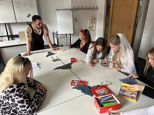 Teilnehmende in Halloween-Kostümen sitzen an Gruppentischen und zeichnen; ein Dozent mit Facepaint lehnt am Tisch, auf dem Fledermaus-Deko liegt.