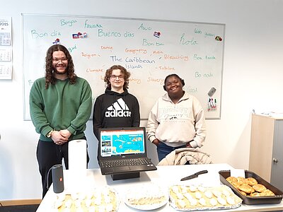 Drei Schüler*innen stehen vor einem Whiteboard, auf dem in der Mitte "The Caribbean Islands" geschrieben steht und außen herum Begriffe in verschiedenen Sprachen. Auf dem Tisch vor der Schülergruppe befindet sich ein Laptop, auf dessen Bildschirm eine Landkarte der karibischen Inseln zu sehen ist. Davor kann man verschiedene Kuchen und Gebäck erkennen.
