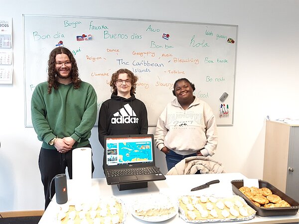 Der nächste Schritt zur Karriere: Career Week Drei Schüler*innen stehen vor einem Whiteboard, auf dem in der Mitte "The Caribbean Islands" geschrieben steht und außen herum Begriffe in verschiedenen Sprachen. Auf dem Tisch vor der Schülergruppe befindet sich ein Laptop, auf dessen Bildschirm eine Landkarte der karibischen Inseln zu sehen ist. Davor kann man verschiedene Kuchen und Gebäck erkennen.