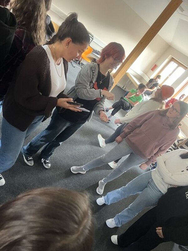 Gutgelaunter gemeinsamer Start ins Schuljahr Junge Erwachsene stehen in einem Kreis in einem Raum zusammen. Eine Person hat ein Smartphone in der Hand.