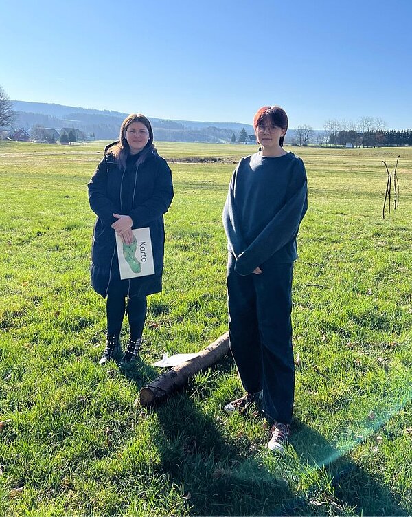 Zwei junge Erwachsene stehen auf einer weitläufigen Wiese mit Blick auf eine hügelige Landschaft im Hintergrund. Die linke Person hält ein Blatt mit einer Karte in der Hand, auf dem das Wort „Karte“ steht. Neben ihnen liegt ein Ast am Boden. Es ist sonnig und klar, die Stimmung wirkt ruhig und konzentriert.