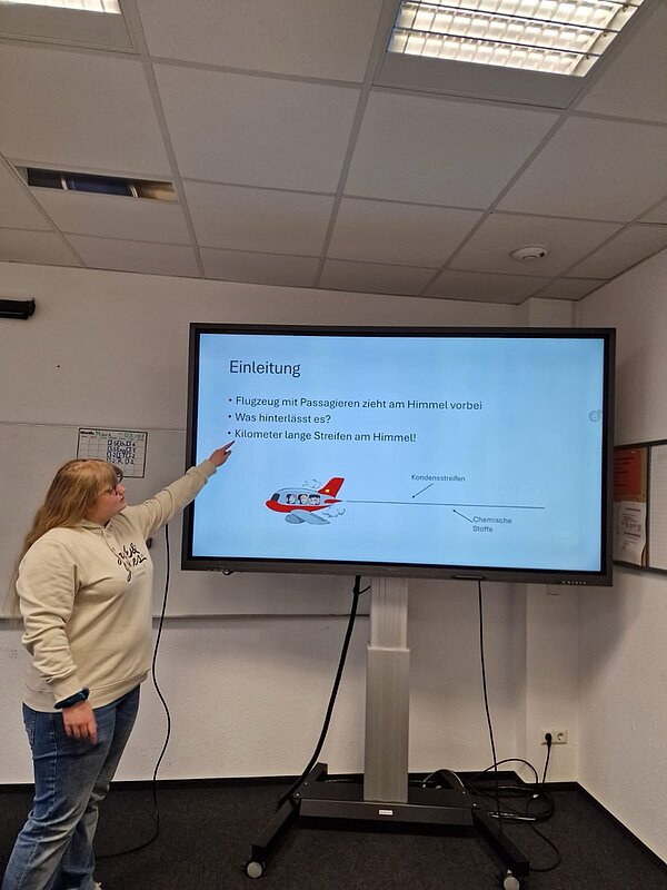 Eine Schülerin steht vor einem Bildschirm und zeigt auf eine Präsentationsfolie mit dem Titel „Einleitung“. Die Folie thematisiert Kondensstreifen von Flugzeugen und mögliche chemische Stoffe, die dabei freigesetzt werden. Ein illustriertes Flugzeug mit Passagieren ist ebenfalls zu sehen.