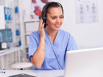 Eine Frau mit blauem Kittel sitz vor einem Computer und hat dabei ein Headset auf.