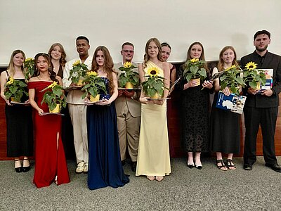 Eine Gruppe junger Menschen in Abendkleidern. Sie halten Zeugnismappen und Töpfe mit Sonnenblumen.