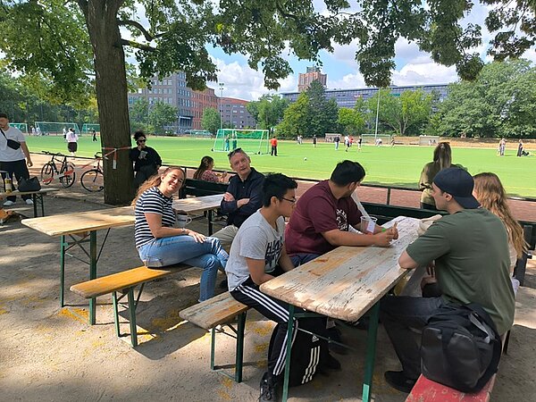 Schnitzeljagd, Tombola und Volleyball-Turnier Schülerinnen und Schüler sowie Lehrkräfte sitzen auf Holzbänken an Tischen am Rand des Sportplatzes und beobachten die Aktivitäten.