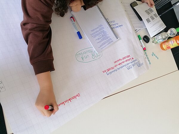 Projekt „Peer-Ausbildung: Schulkino“ durch Alter Gasometer e.V. Zwickau Person schreibt mit rotem Stift auf ein großes Plakatpapier, umgeben von Notizen, Stiften und Getränken. Das Thema bezieht sich auf NSU und Hintergründe.