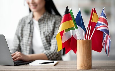 Eine Frau mit Headset sitzt an einem Schreibtisch und tippt auf einem Laptop. Neben dem Laptop liegen ein Block und ein Stift. Auf dem Schreibtisch, im rechten Rand des Bildes steht eine Holzvase, in der verschiedene europäische Flaggen stecken.