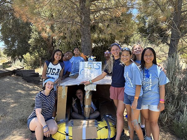 Eine Gruppe junger Menschen steht und sitzt um eine selbstgebaute Konstruktion aus Pappe und gelben Luftmatratzen, die unter einem Baum im Freien platziert ist, im Rahmen eines Campaufenthaltes. Die Konstruktion hat ein Schild mit der Aufschrift 'LET'S GO'. Die Jugendlichen tragen sommerliche Kleidung, darunter T-Shirts und Shorts, und einige haben Armbänder an den Handgelenken. Im Hintergrund sind weitere Bäume und ein sonniger Himmel zu sehen. Die Szene vermittelt eine entspannte Atmosphäre während eines Auslandsaufenthaltes einer Auszubildenden der Euro Akademie Bamberg in San Diego.