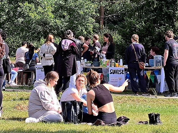 Auf einer Wiese sitzen drei Frauen, die rechte zeigt mit ihrer Hand auf einen Punkt außerhalb des Bildes. Im Hintergrund stehen viele Menschen an Tischen, auf denen ein Buffet aufgebaut ist und Getränke ausgeschenkt werden.
