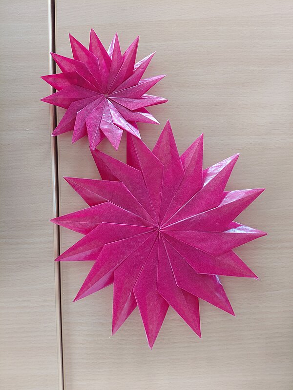 Zwei pinkfarbene, sternförmige Papierfaltungen in unterschiedlicher Größe hängen dekorativ an einer hellen Holztür.