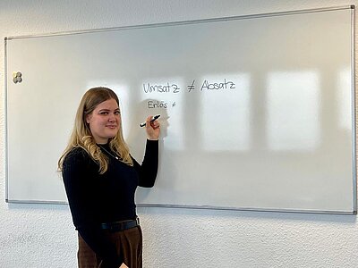 Die Lehrerin Anna Eschenbach steht in dunkler Kleidung vor einem Whiteboard, auf das sie die Wörter "Umsatz ist ungleich Absatz" und "Erlös ist ungleich …" geschrieben hat. Der zweite Vergleich ist (noch) nicht vollständig.