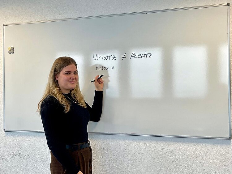 Die Lehrerin Anna Eschenbach steht in dunkler Kleidung vor einem Whiteboard, auf das sie die Wörter "Umsatz ist ungleich Absatz" und "Erlös ist ungleich …" geschrieben hat. Der zweite Vergleich ist (noch) nicht vollständig.