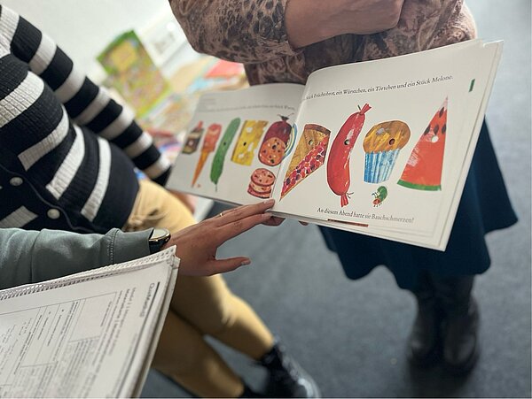 Mathe im Kindergarten? Geht und macht Spaß! Drei Schülerinnen schauen sich eine Seite des Buchs "Die kleine Raupe Nimmersatt" an, man sieht aber keine Gesichter der Personen.