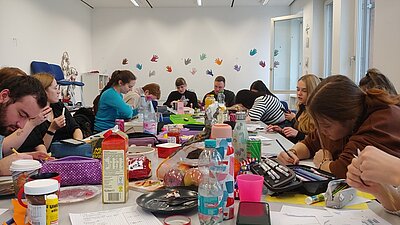 Schülerinnen und Schüler sitzen an langem Tisch, mit zahlreichen Bastelutensilien und sind vertieft in ihre kreativen Arbeiten.