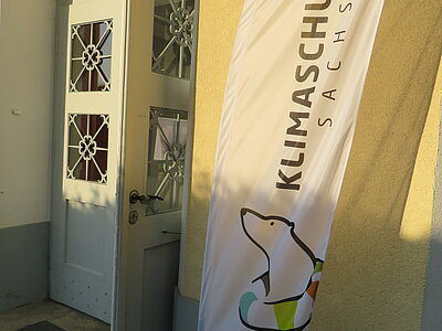 Vor einer gelben Wand und einer weißen Tür steht eine Fahne mit einem Eisbären und der Aufschrift "Klimaschule Sachsen".