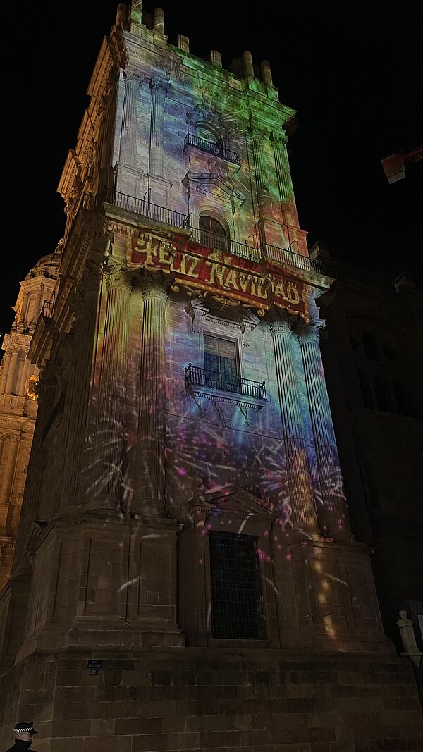 Historische Kirchenfassade, die nachts bunt mit Lichteffekten und dem Schriftzug „Feliz Navidad“ angestrahlt wird, wodurch Säulen und Mauern in kräftigen Farben leuchten.