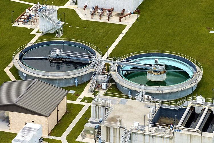 Zwei große, kreisförmige Becken mit Metallbrücken und technischen Aufbauten zur Abwasseraufbereitung in einer Kläranlage stehen nebeneinander auf einer gepflegten Grünfläche. Die Becken sind von Wegen und weiteren Gebäuden umgeben. In einem Becken ist dunkles Wasser zu sehen, im anderen helles Wasser. Mehrere Rohrleitungen und technische Anlagen befinden sich im Hintergrund.