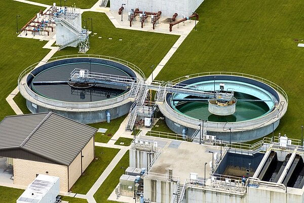 Zwei große, kreisförmige Becken mit Metallbrücken und technischen Aufbauten zur Abwasseraufbereitung in einer Kläranlage stehen nebeneinander auf einer gepflegten Grünfläche. Die Becken sind von Wegen und weiteren Gebäuden umgeben. In einem Becken ist dunkles Wasser zu sehen, im anderen helles Wasser. Mehrere Rohrleitungen und technische Anlagen befinden sich im Hintergrund.