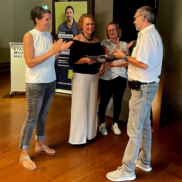 Drei Lehrerinnen und ein Lehrer stehen lächelnd zusammen. Der Lehrer überreicht der Absolventin ihr Abschlusszeugnis und eine Rose.