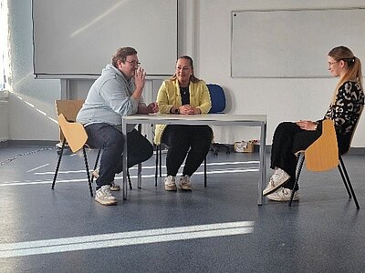 In einem Klassenzimmer sitzen zwei Frauen und ein Mann auf Stühlen an einem Tisch