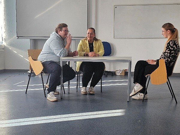 In einem Klassenzimmer sitzen zwei Frauen und ein Mann auf Stühlen an einem Tisch