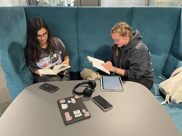 Zwei Personen sitzen in einer blauen Sitznische und lesen Bücher. Auf dem runden Tisch vor ihnen liegen Tablet, Smartphone, MP3-Player, Kopfhörer und ein Laptop; im Hintergrund große Fenster.